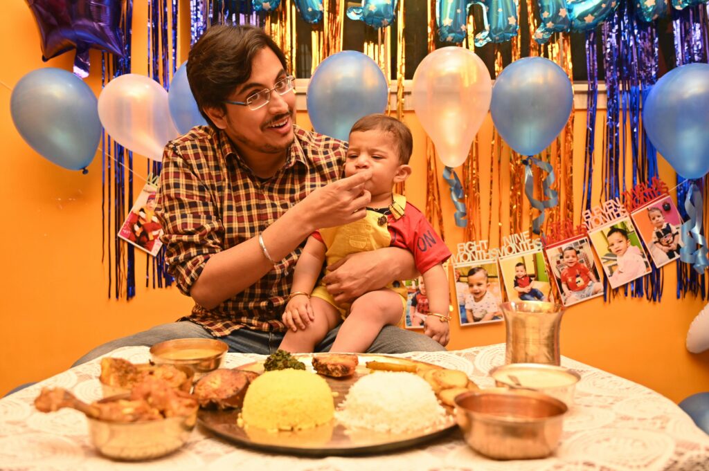 A joyful father feeds his baby son during a vibrant Indian birthday celebration with traditional food and decorations.